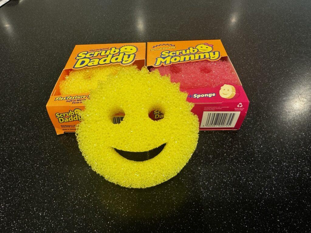 Bildet viser en gul, smilende skuresvamp (Scrub Daddy) plassert foran to esker: en oransje Scrub Daddy-eske og en rosa Scrub Mommy-eske. Den gule svampen har et ansikt med øyne og en smilende munn, og den står på en mørk overflate med små hvite prikker. De fargerike eskene i bakgrunnen har logoene og bilder av svampene, som matcher svampens design og farger.