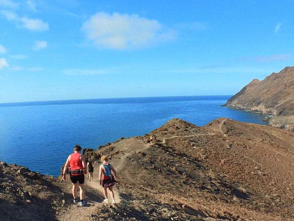 Bildet viser to personer som går langs en smal sti på en steinete ås eller fjellrygg med utsikt mot havet. Personen i forgrunnen, iført en rød topp og shorts, bærer en liten ryggsekk, mens den andre personen følger like bak. Landskapet rundt er tørt og steinete, typisk for et karrig kystområde. Stien snor seg langs åssiden, og i bakgrunnen kan man se det klare, blå havet som strekker seg ut mot horisonten under en lys blå himmel med noen spredte skyer. Bildet formidler en følelse av utforskning og eventyr i et dramatisk, men fredelig kystmiljø. Andreas Gossner fra Løpetrening.no på treningsreiser og løpereiser på Fuerteventura (kanariøyene) med en av flere grupper høst- eller vinterstid.