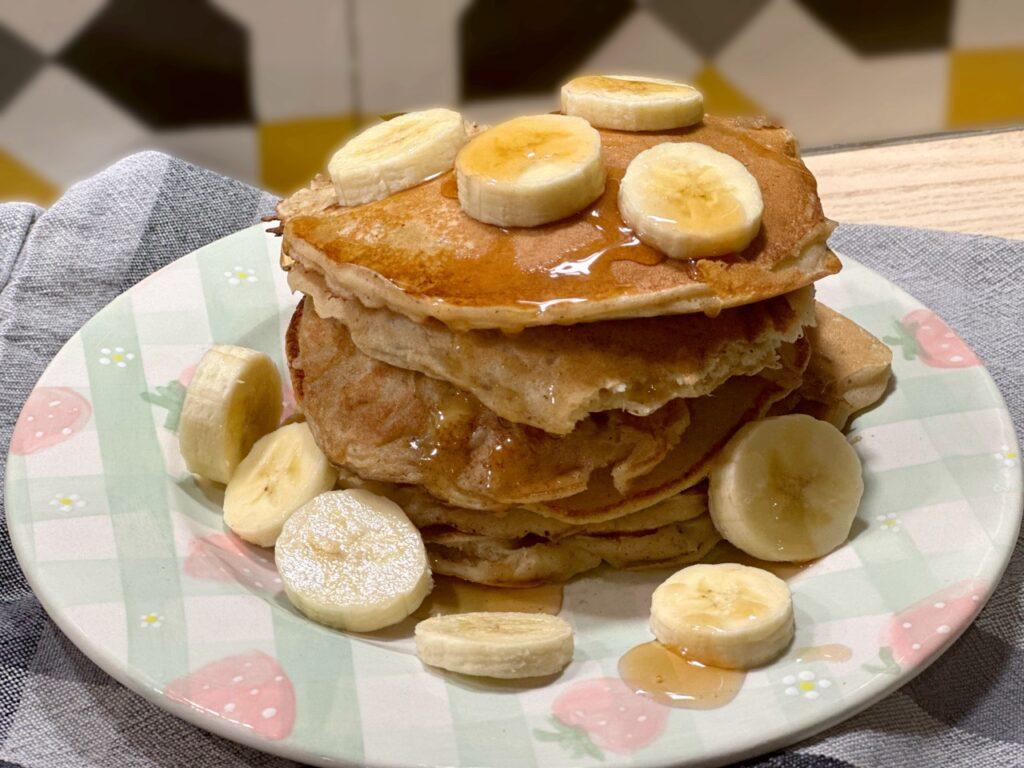 Bildet viser en fristende stabel av gyllenbrune bananpannekaker, toppet med skiver av fersk banan og drysset med honning eller lønnesirup. Pannekakene er tykke og luftige, servert på en dekorativ tallerken med et lyst mønster. Rundt stabelen ligger flere bananskiver som komplementerer retten, og alt dette står på en grå tekstil serviett.