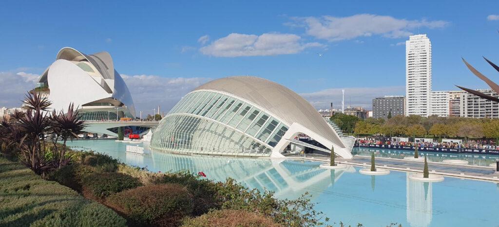 Bildet viser et moderne og futuristisk arkitektonisk kompleks ved en vannspeil i Valencia, Spania. Den store, hvite strukturen i forgrunnen ser ut som Hemisfèric, en del av Ciutat de les Arts i les Ciències (Kunst- og vitenskapsbyen). Bygningen har en karakteristisk kuppelform, som ligner på et gigantisk øye. I bakgrunnen kan man se en annen modernistisk bygning, Palau de les Arts Reina Sofia, som fungerer som et operahus. Vegetasjonen rundt vannspeilet gir et grønt preg til området, mens vannet reflekterer den futuristiske arkitekturen. Det er også skilt med teksten "Heineken" på venstre side, som indikerer at dette bildet ble tatt i et arrangement, kanskje Valencia Marathon, med et målområde som er synlig langs vannet. Løpetrener og løpeekspert, Andreas Gossner, forteller om sin egen treningsreise mot maraton i Valencia i desember 2024!