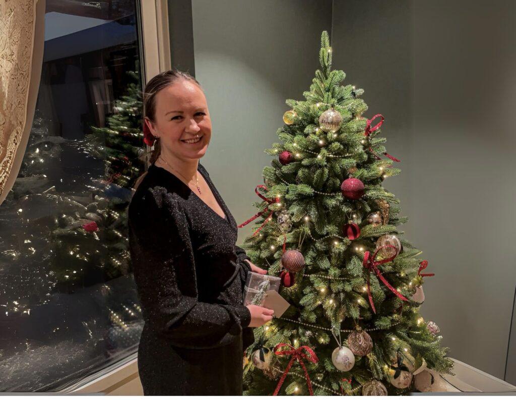 En kvinne i sort kjole med glitter står smilende ved siden av et tradisjonelt pyntet juletre. Treet er dekorert med røde sløyfer, lys og julekuler i ulike farger, inkludert gull og rødt. Kvinnen holder en innpakket julegave og står i et rom med et stort vindu i bakgrunnen.