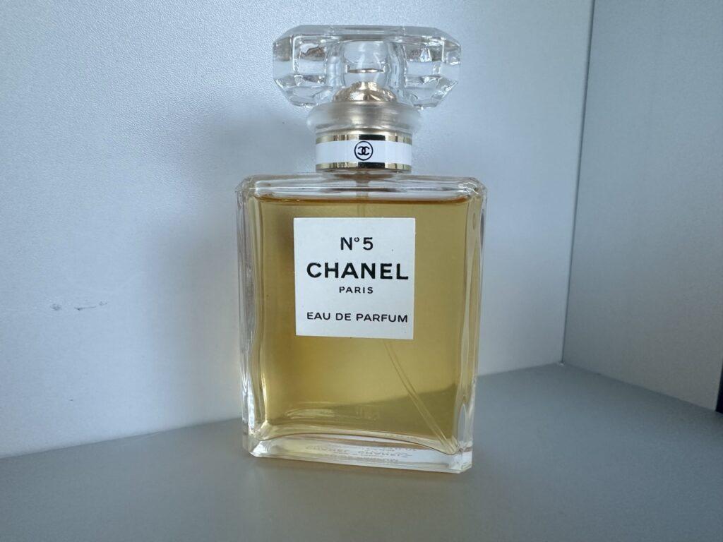 En klar glassflaske med den velkjente parfymen Chanel No. 5. Flasken har et minimalistisk design med en firkantet form, fylt med en gylden væske. På etiketten står det 'CHANEL No. 5 Paris Eau de Parfum.' Flasken står plassert mot en enkel, lys bakgrunn som fremhever dens klassiske og stilrene utseende.