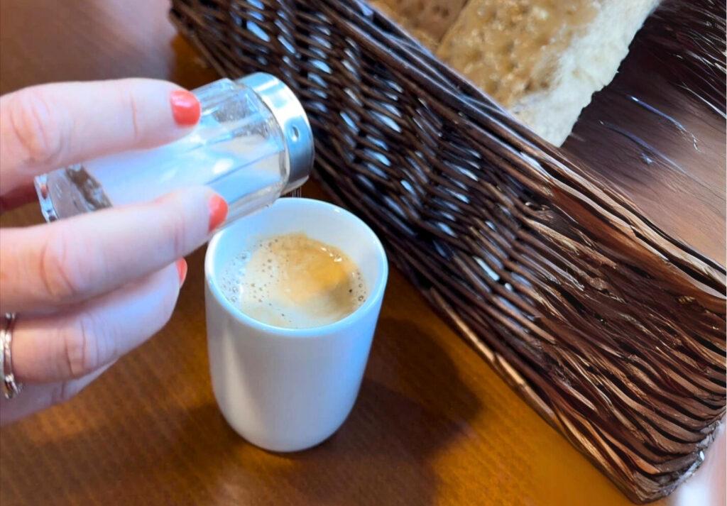 En nærbilde viser en hånd med røde negler som drysser salt fra en saltbøsse ned i en kopp med kaffe. Kaffen har et lett skumlag på toppen. Ved siden av står en kurv med brød, og overflaten er et mørkt trebord som gir en varm atmosfære.