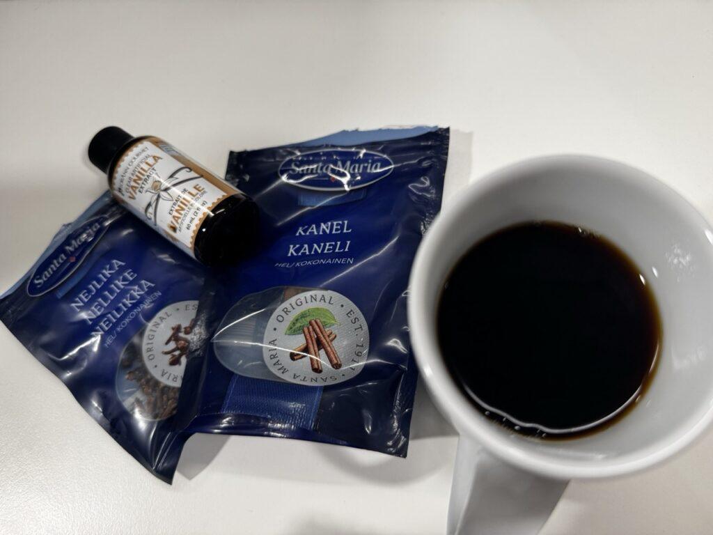 En kopp svart kaffe står ved siden av krydderposer med kanel og nellik, samt en flaske vaniljeekstrakt. Dette er ingredienser som kan brukes for å gi kaffen en ekstra smaksopplevelse