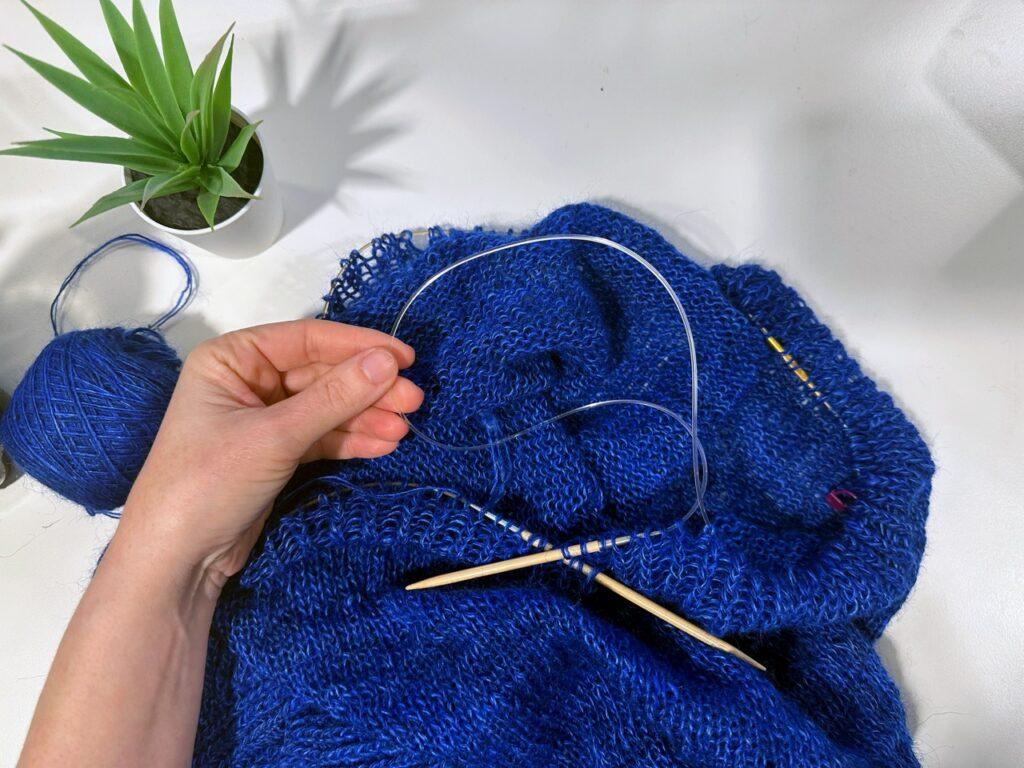 Bildet viser en hånd som holder en strikkepinne og strikkearbeid i dyp blå farge, laget med teknikken "traveling loop." Strikketøyet ligger på en lys flate ved siden av et grønt plante i potte. Garnnøstet er blått, og det brukes rundpinner i tre.