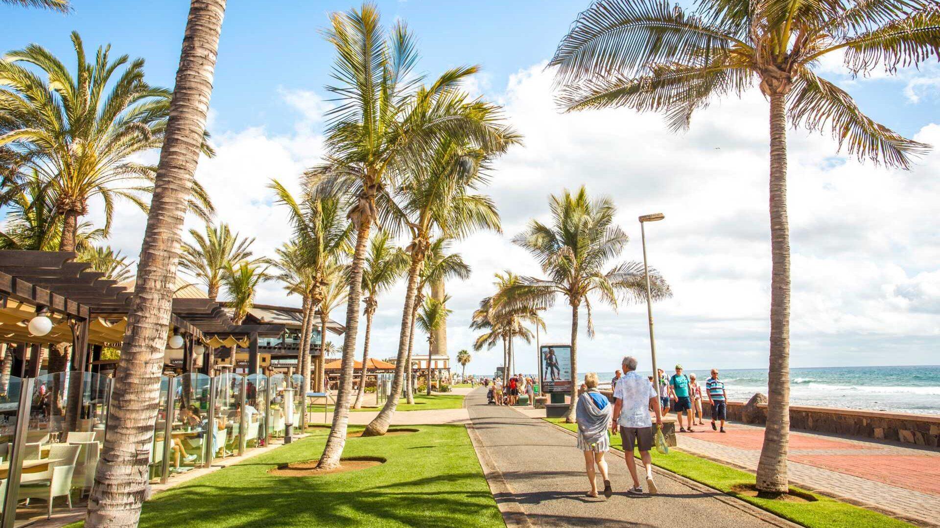 Dette bildet viser en promenade langs kysten på Gran Canaria, som kan være et fristende reisemål for en vinterreise. Palmetrær med slanke stammer står på rekke og rad langs en bred gangvei. Solen skinner, og himmelen er lyseblå med noen få hvite skyer, som skaper en lys og varm atmosfære. Langs venstre side er det uteserveringer med glassvegger og bord hvor folk kan sitte og nyte måltider. På høyre side ser vi havet som strekker seg utover med små bølger som bryter mot stranden. Flere mennesker spaserer langs promenaden, kledd i lette sommerklær, noe som gir en avslappet og feriepreget stemning. Dette gir en sterk kontrast til vinterens kalde klima og gjør Gran Canaria til et populært tilfluktssted for de som søker sol og varme.