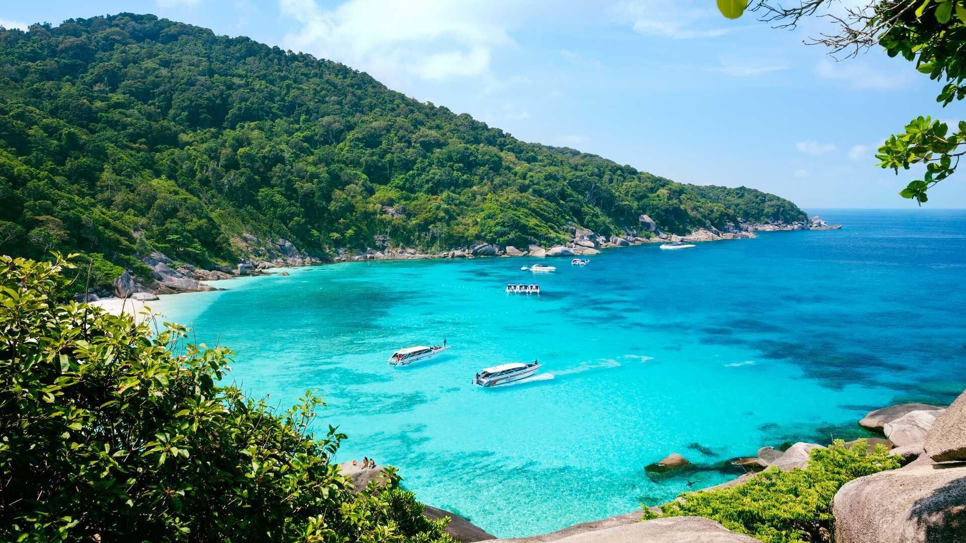 Dette bildet viser en nydelig bukt i Thailand, et ideelt reisemål for en vinterreise til sol og varme. Bukten er fylt med krystallklart, turkisblått vann som glitrer under solen. I bakgrunnen strekker en frodig, grønnkledd åsside seg opp fra kysten, med tett, tropisk vegetasjon som dekker hele området. Flere små båter ligger forankret i bukten, klare til å ta besøkende på eventyr, kanskje til snorkling eller utforsking av de nærliggende strendene. Bildet oser av eksotisk skjønnhet og fred, med klare, solrike himler som gir en følelse av å være langt unna vinterens kulde. Dette gjør Thailand til en perfekt flukt fra den kalde vinteren, hvor man kan nyte tropiske omgivelser og varme temperaturer.