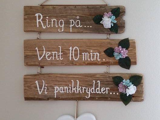 Skilt laget av drivved med skriften "Ring på... Vent 10 min. Vi panikkrydder". Pyntet med blomster og hjerte.