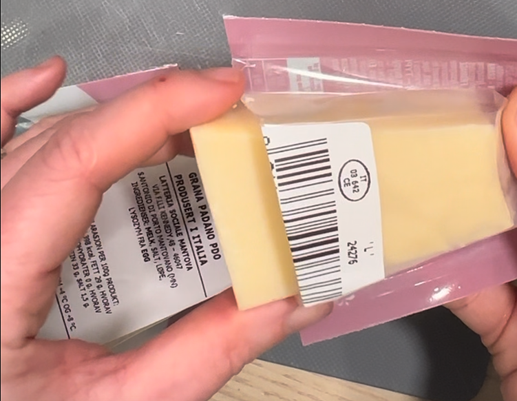 Nærbilde av to hender som holder en bit Grana Padano i original emballasje. Osten er delvis pakket ut, og etiketten med produktinformasjon og strekkode er tydelig synlig. Bakgrunnen består av en grå matte og et lyst trebord, som skaper en nøytral og funksjonell setting. Bildet illustrerer praktisk bruk av emballasje for oppbevaring av ost.