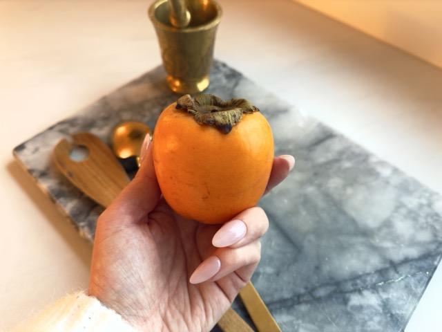 n person holder en kaki (persimmon) i hånden over en marmorbordplate. Gyldent bestikk og en morter i messing er synlige i bakgrunnen, og fruktens intense oransje farge står i sentrum.