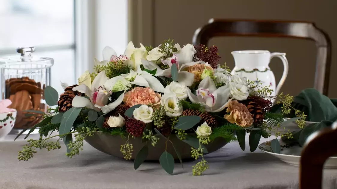 Bildet viser en vakker juleinspirert blomsterdekorasjon plassert på et dekket bord. Dekorasjonen består av hvite orkidéer, roser, nellik og grønne blader, supplert med kongler og røde detaljer som gir et klassisk og elegant preg. I bakgrunnen kan man se julepyntet servise, inkludert en bolle med julesnacks og en krukke med pepperkaker. Hele oppsettet gir en varm og koselig julestemning, perfekt for en høytidelig middag eller som en dekorativ gave.