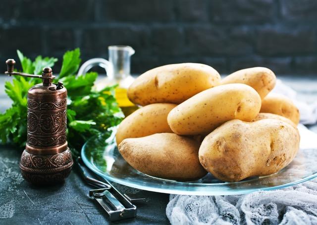 En stabel med gyldne poteter plassert på en glassplate. Potetene er omgitt av fersk persille, en flaske olivenolje og en dekorativ krydderkvern i kobber. Den mørke bakgrunnen fremhever ingrediensene og gir en sofistikert, rustikk atmosfære.