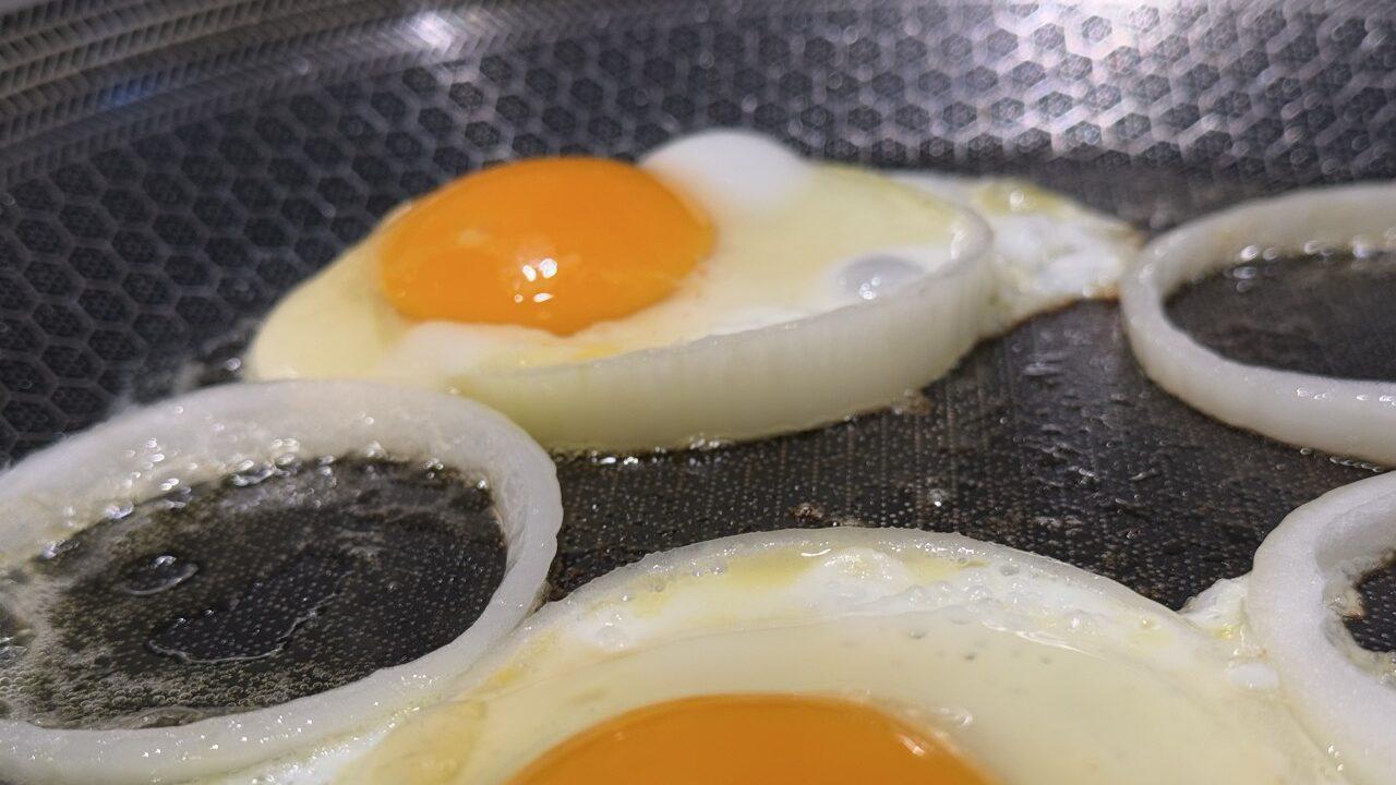 Nærbilde av stekte egg som tilberedes i ringer av løk på en stekepanne med bikubemønstret non-stick-overflate. Eggeplommene har en klar, gylden oransje farge, og eggehviten har stivnet jevnt rundt løkringene. Pannen har en lett oljet overflate som glinser under lyset, og detaljene i strukturen på stekepannen kommer tydelig frem. Bildet gir et appetittvekkende og moderne inntrykk av en kreativ matrett.