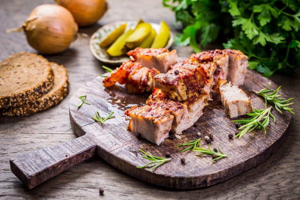 Et rustikt serveringsbrett med saftige biter av sprøstekt ribbe, dandert med ferske rosmarinkvister og krydderkorn. I bakgrunnen skimtes skiver av grovt brød, sylteagurk og løk, mens frisk persille gir en grønn kontrast. Ribben har en gyllen, sprø svor og et saftig indre, perfekt til tradisjonelle måltider.