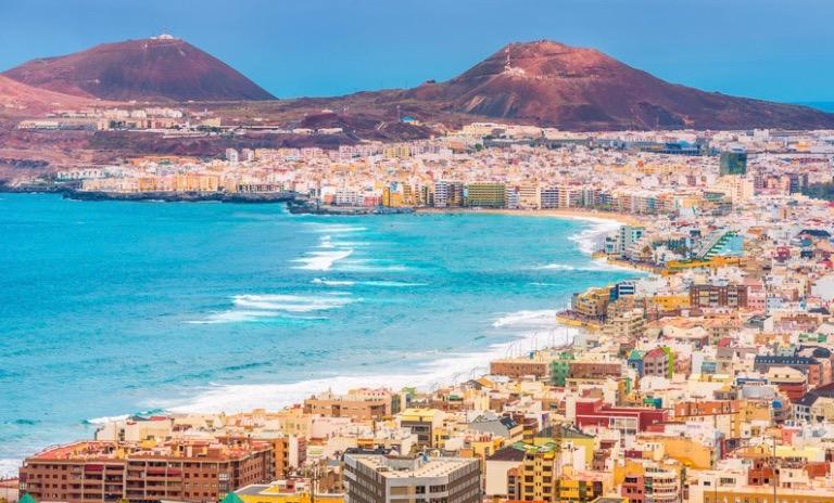 Bildet viser en vakker kystby ved havet, trolig Las Palmas på Gran Canaria. Forgrunnen er fylt med bygninger i ulike farger og arkitektoniske stiler. Langs vannkanten kan man se en strand som strekker seg langs byen. Havet er klart og blått, med bølger som ruller inn mot land. I bakgrunnen er det fjell, som gir en dramatisk kontrast til kystlandskapet. Stemningen er livlig og typisk for et solfylt, turistvennlig sted ved Atlanterhavet. kanariøyene er en stor favoritt blant rensende nordmen og mange legger vinterfeien, juleferie og høstferie hit.