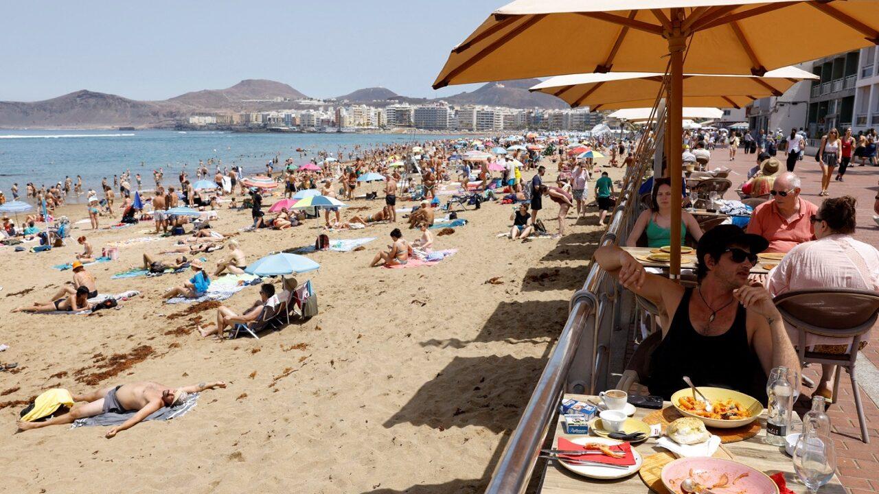 Bildet viser en solrik strand på Gran Canaria, full av turister som nyter det varme været. I forgrunnen sitter flere personer ved et bord på en restaurant med parasoller, mens de spiser og slapper av. Bak dem er stranden full av solbadere og parasoller, med noen som ligger på sanden og andre som står og går. Langs stranden sees moderne bygninger og fjell i bakgrunnen, som setter den vakre kysten i kontrast.