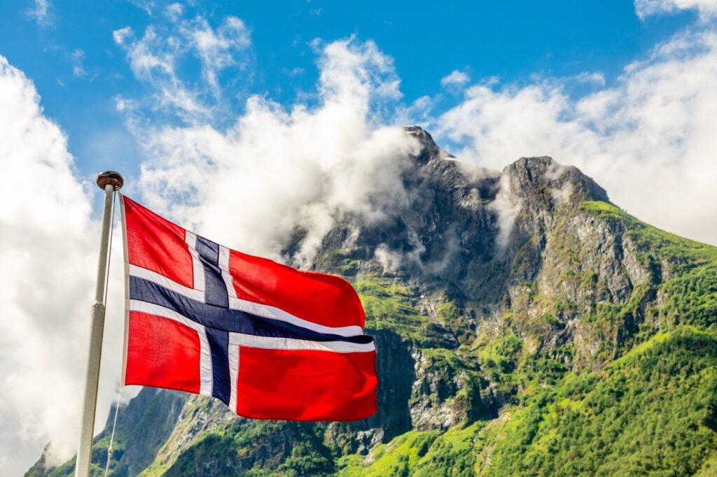 Bildet viser et norsk flagg som vaier i vinden, med en vakker bakgrunn av majestetiske fjell og frodig grønn vegetasjon under en blå himmel med lette, hvite skyer. Flagget er i klare farger, rødt med hvitt og blått kors, og symboliserer nasjonal stolthet. Fjellene i bakgrunnen skaper en dramatisk og nasjonalromantisk stemning, som ofte forbindes med Norge. Dette bildet kan brukes som en visuell innledning til en artikkel om røde dager og fridager i Norge i 2025. Her er viktige dager å inkludere i teksten: 1. januar (Nyttårsdag): En nasjonal fridag for å markere starten på det nye året. Påske (24.–28. mars): Skjærtorsdag (20. mars), langfredag (21. mars), påskedag (24. mars), og andre påskedag (25. mars) er røde dager. 1. mai (Arbeidernes dag): En dag for arbeidernes rettigheter og en fridag. 17. mai (Grunnlovsdagen): Norges nasjonaldag med feiringer over hele landet. Kristi himmelfartsdag (29. mai): En viktig kristen helligdag. Pinse (8.–9. juni): Første pinsedag (8. juni) og andre pinsedag (9. juni) er fridager. 25. desember (1. juledag) og 26. desember (2. juledag): Viktige dager i julen. Artikkelen kan også gi tips om hvordan man kan planlegge ferie for å få lengre sammenhengende fri i 2025 ved å kombinere røde dager med inneklemte dager eller helger.