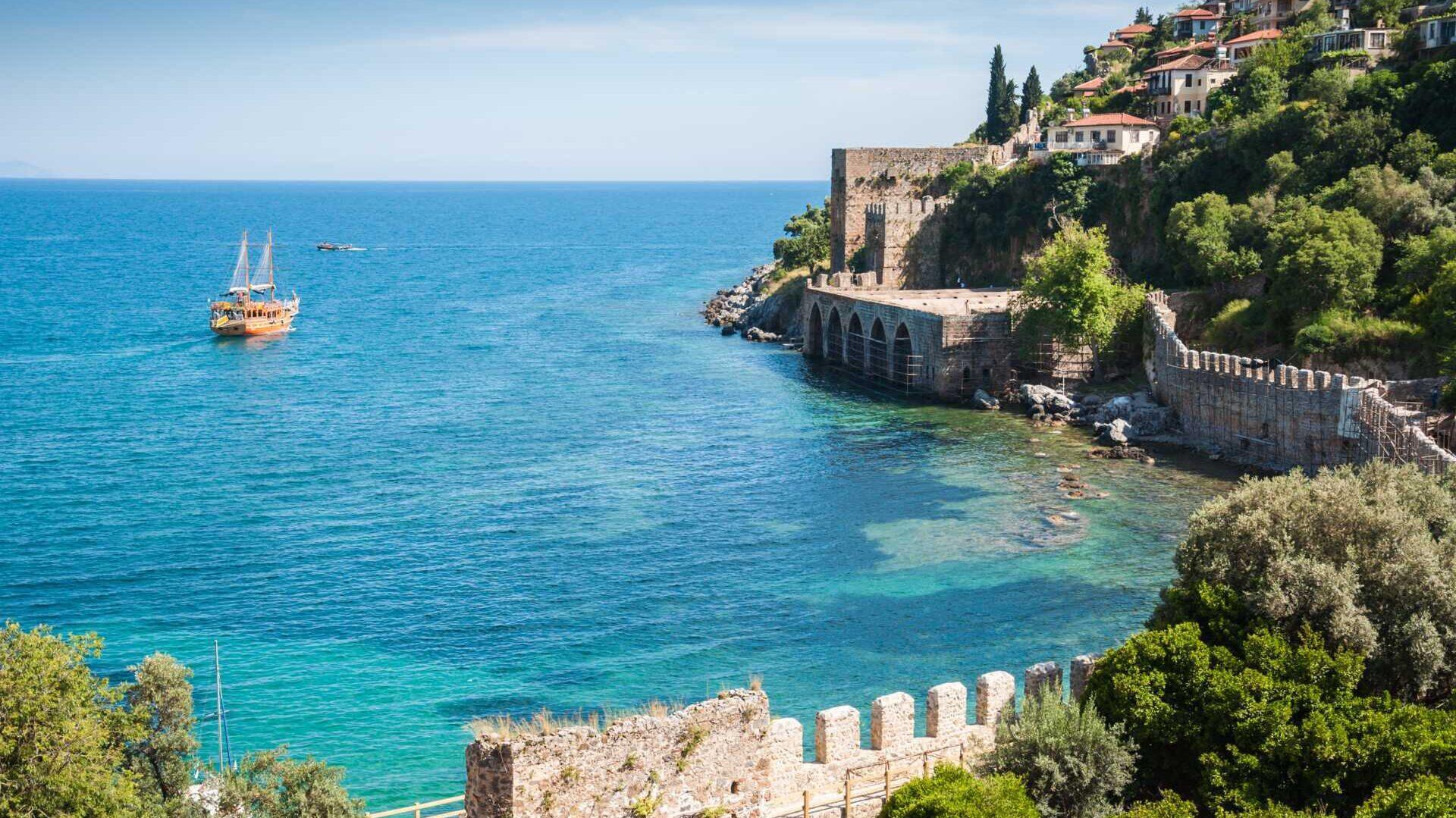 Bildet viser den ikoniske Hagia Sophia i Istanbul, Tyrkia, med en stor, blomsterdekorert park og en fontene i forgrunnen. Hagia Sophia er en imponerende bygning med kupler og minareter, omgitt av frodige trær og fargerike blomsterbed som skaper en vakker og rolig atmosfære. Mange mennesker spaserer og nyter området i det solfylte været.