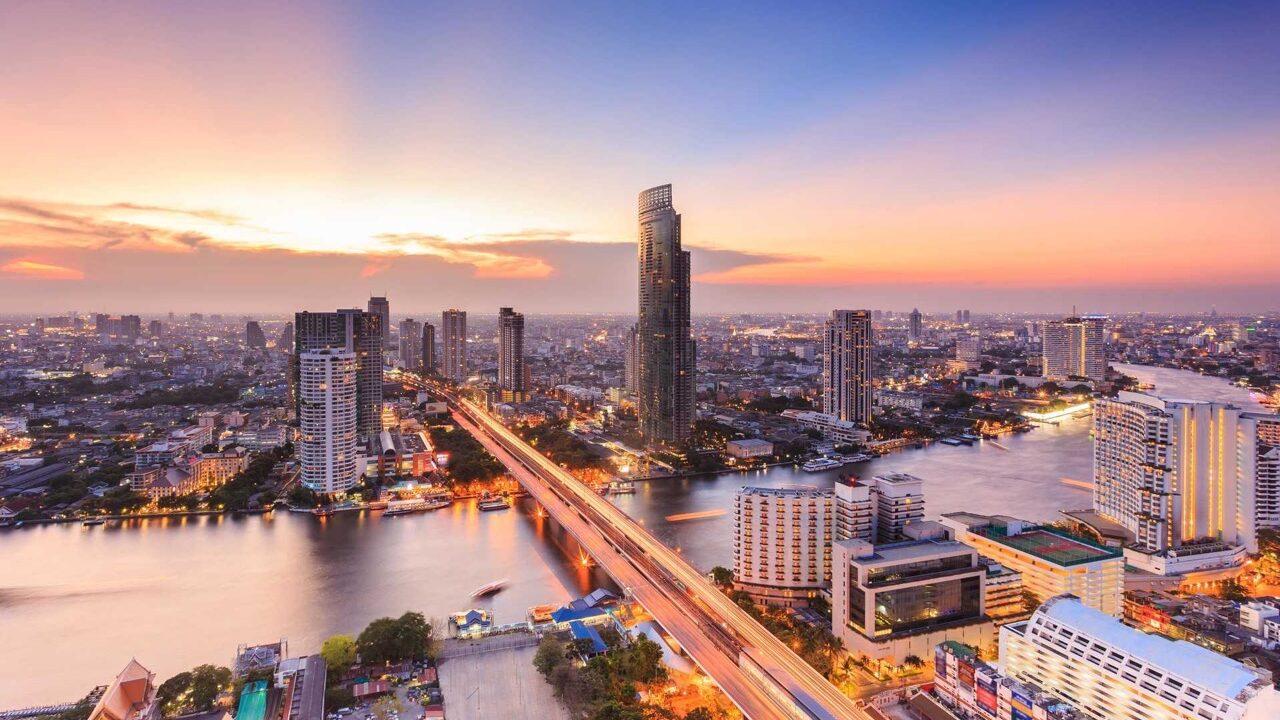 Bildet viser en spektakulær solnedgang over Bangkok, Thailands pulserende hovedstad. Chao Phraya-elven slynger seg gjennom byen, omkranset av skyskrapere, broer og lysende bygninger. Den travle metropolen kommer til live med en blanding av moderne arkitektur og tradisjonelle elementer.