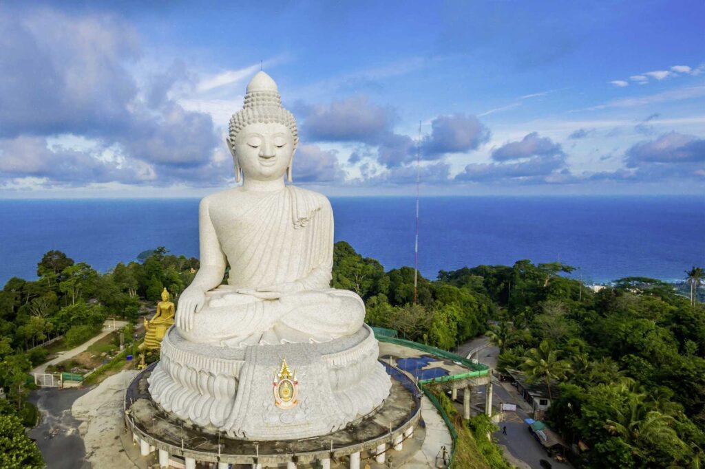 Bildet viser den majestetiske Big Buddha-statuen i Phuket, en av øyas mest ikoniske attraksjoner. Den 45 meter høye hvite marmorstatue sitter i en rolig meditasjonsstilling på toppen av en grønnkledd høyde, med panoramautsikt over havet og det frodige landskapet. Skyene svever over en blå himmel, og en mindre gylden Buddha-statue kan sees i bakgrunnen, noe som gir stedet en spirituell og fredelig atmosfære.