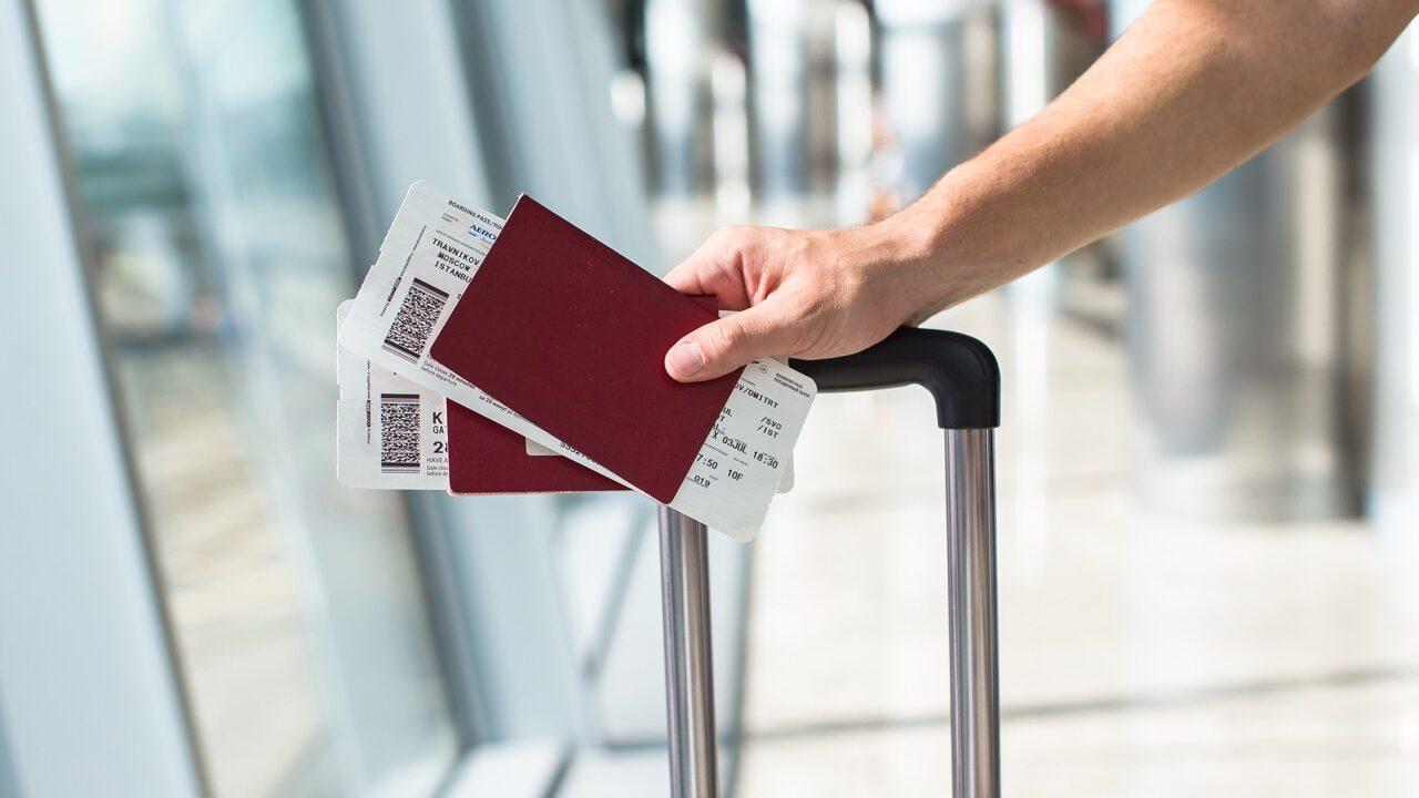 Bildet viser en person som holder et pass og flere boardingkort i hånden, plassert oppå et koffert-håndtak. Settingen er en flyplass, noe som indikeres av de store glassvinduene i bakgrunnen. Bildet symboliserer reiselyst og praktiske reisedetaljer, samtidig som det kan illustrere temaer som flybilletter, passkrav og forberedelser til reisen. Det er et ideelt bilde for å understøtte en artikkel om flyreiser eller praktiske tips for å spare penger på flybilletter.