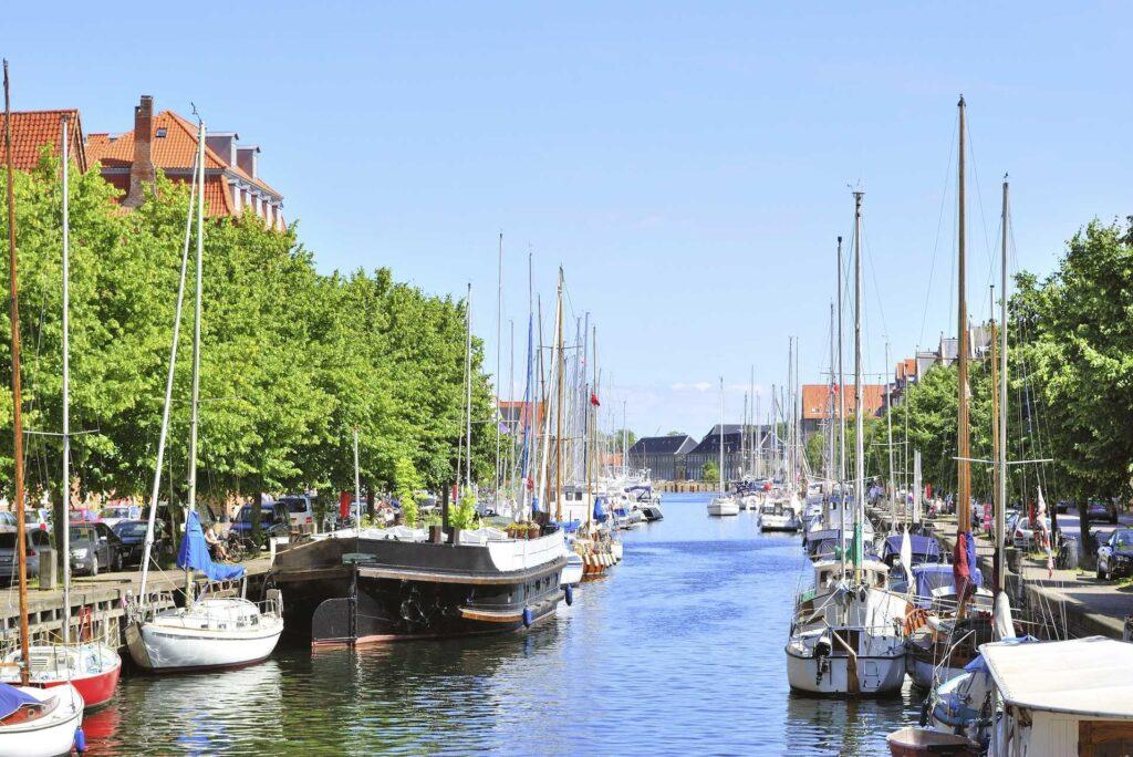 Bildet viser en idyllisk kanal omgitt av grønne trær og båter som ligger til kai. Kanalens speilblanke vann reflekterer mastene på seilbåtene, og langs kanalbredden er det murhus med røde tak. Himmelen er klar og blå, og atmosfæren virker rolig og sommerlig.