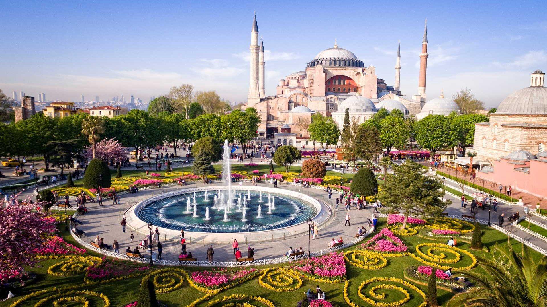 Bildet viser den ikoniske Hagia Sophia i Istanbul, Tyrkia, med en stor, blomsterdekorert park og en fontene i forgrunnen. Hagia Sophia er en imponerende bygning med kupler og minareter, omgitt av frodige trær og fargerike blomsterbed som skaper en vakker og rolig atmosfære. Mange mennesker spaserer og nyter området i det solfylte været.