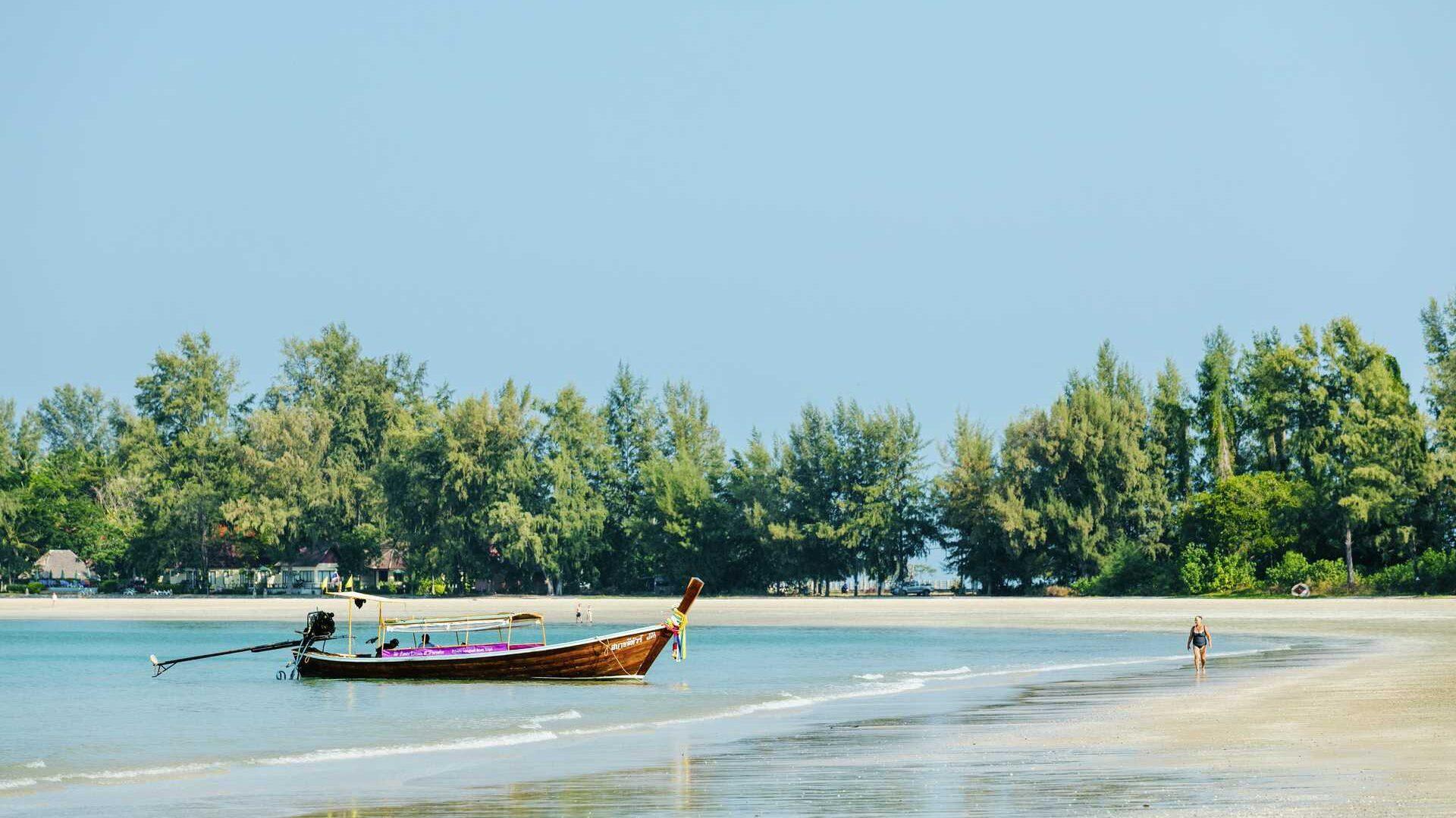 Bildet viser en fredelig, uberørt strand i Thailand med myk, gyllen sand og en tradisjonell longtail-båt som ligger forankret i det klare, blå vannet. I bakgrunnen ruver frodige trær langs kysten, mens en enslig strandgjest nyter en rolig spasertur langs vannkanten.