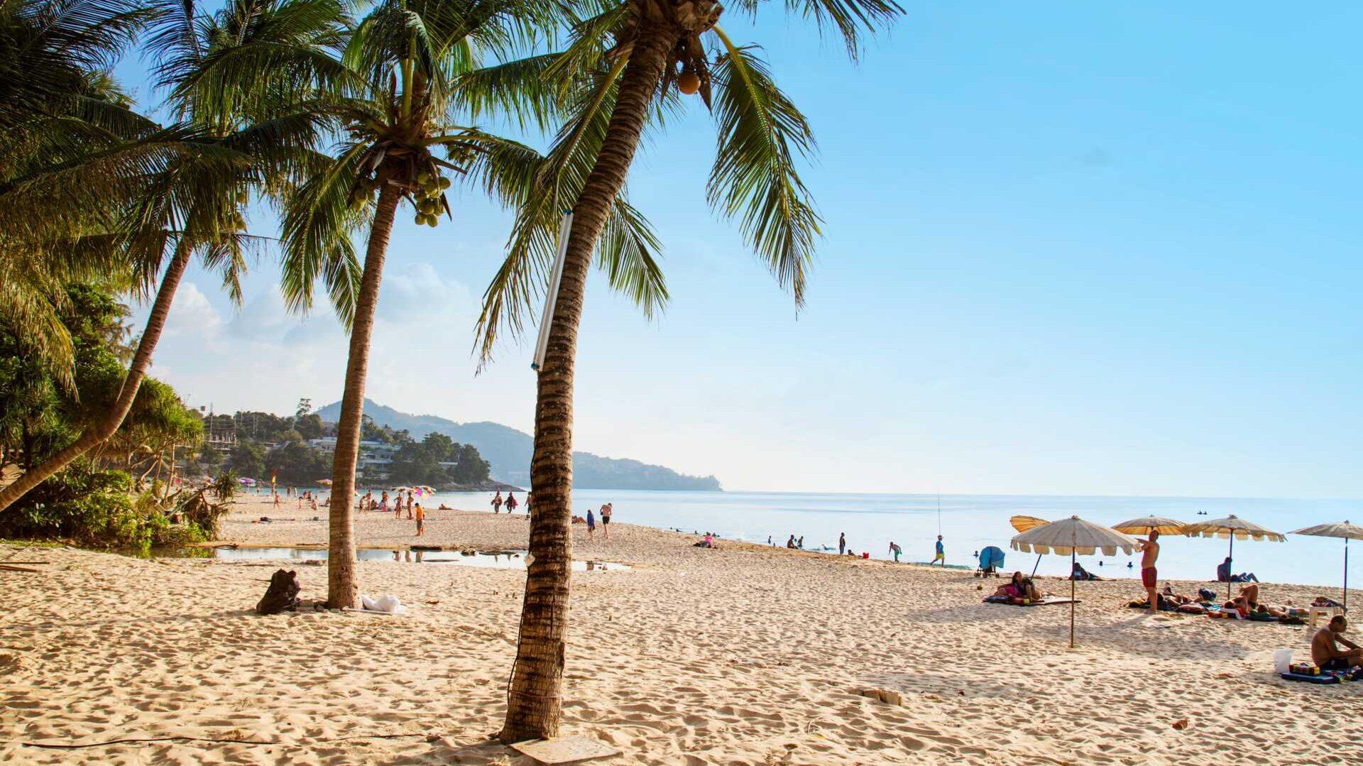 Bildet viser en idyllisk tropisk strand i Thailand med myk, gyllen sand og høye palmetrær som vaier i vinden. Solen skinner fra en skyfri himmel, og strandgjester nyter dagen under parasoller eller bader i det rolige, krystallklare havet.