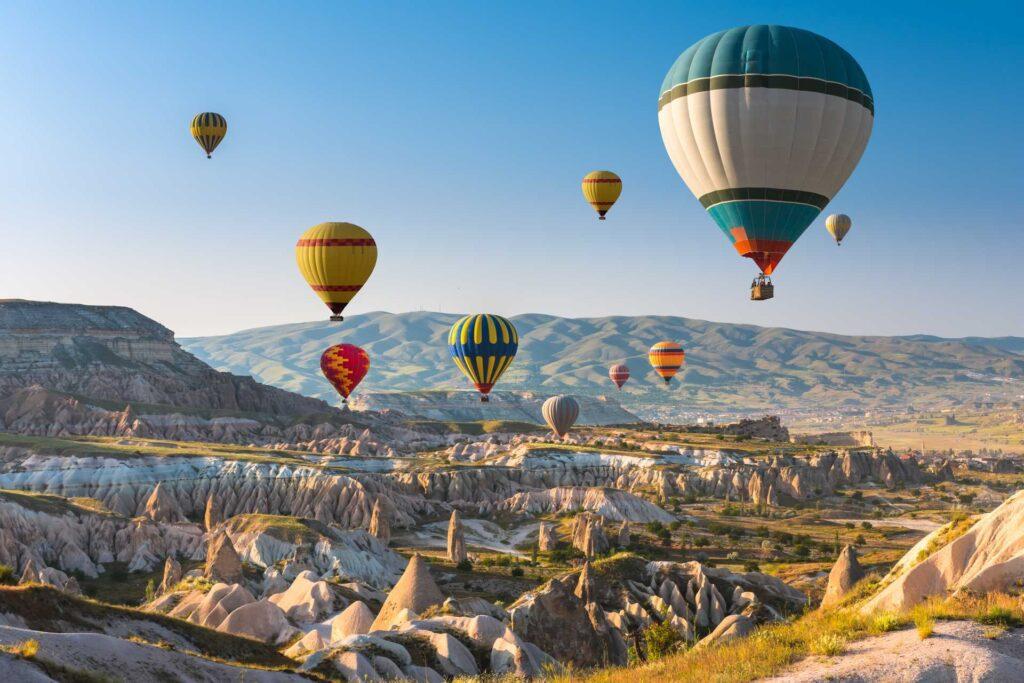 Bildet viser flere fargerike varmluftsballonger som svever over et unikt landskap med steinformasjoner og daler i Cappadocia, Tyrkia. Landskapet består av myke åser, dramatiske klippeformasjoner og grønne sletter, mens himmelen er klar og blå, og ballongene skaper en magisk stemning.