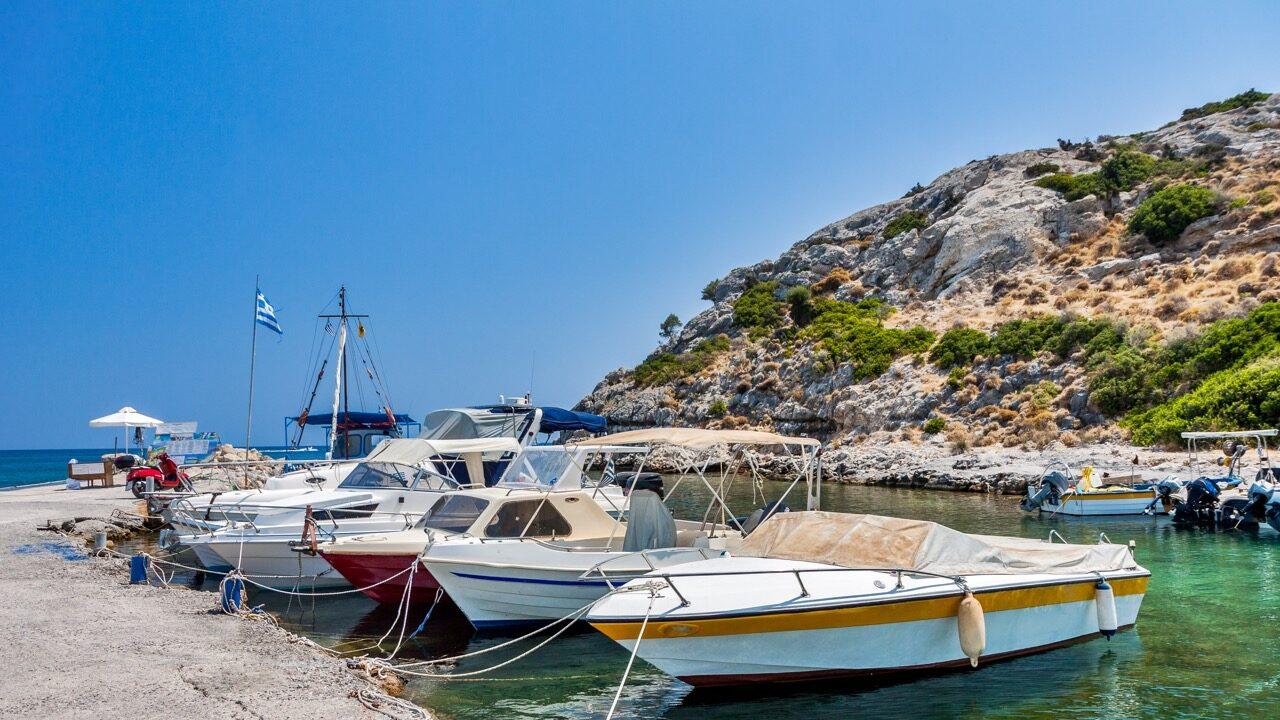 Et bilde av en liten havn på Rhodos, Hellas, med flere småbåter fortøyd ved en brygge. En gresk flagg vaier i vinden ved bryggen, og en person kan sees sittende under en parasoll i bakgrunnen. Området er omgitt av steinete åser med grønn vegetasjon. Vannet er klart og speiler den blå himmelen over. Rhodos i Hellas er en drøm, og her får du både billige reiser og pakkereiser til den greske øya. Vi gir deg også fem ting du må se og gjøre på Rhodos, samt svar på ofte stilte spørsmål om øya.