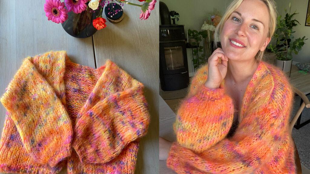 Bildet viser en fargerik, håndstrikket cardigan laget i myk mohair. Til venstre ligger cardiganen brettet på et trebord, omgitt av en vase med friske blomster i sterke farger. Til høyre har en smilende kvinne den på seg, sittende ved et bord i en koselig innredet stue med grønne planter og en vedovn i bakgrunnen. Den fluffy strikkegenseren har varme toner av oransje, rosa og gult, strikket med tykke pinner for et luftig uttrykk.