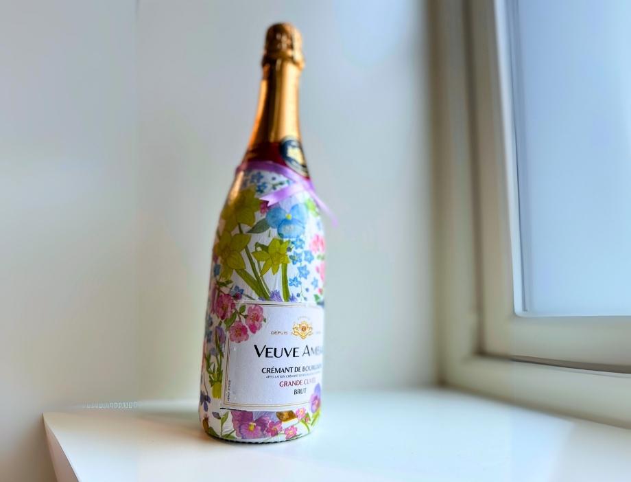 En champagneflaske står på en hvit vinduskarm med en lys bakgrunn. Flasken er dekorert med decoupage-teknikk, hvor et mønstret papir med blomster i blått, gult, rosa og grønt dekker flaskens overflate. Etiketten i midten er synlig, og en tynn lilla sløyfe er knyttet rundt flaskehalsen. Vinduet ved siden av slipper inn naturlig lys, som gir flasken et mykt og delikat uttrykk.