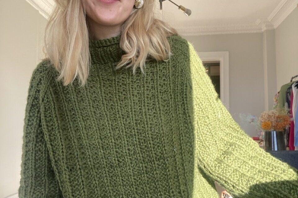 En ung kvinne iført en chunky, strikket genser i en dyp skoggrønn farge. Genseren, kalt Skogdrøm, er fra Vild Knitwear og har en myk, ribbestrikket tekstur med en høy hals og vide ermer. Naturlig dagslys lyser opp plagget, og en koselig innendørs atmosfære skaper en varm og stilfull setting.