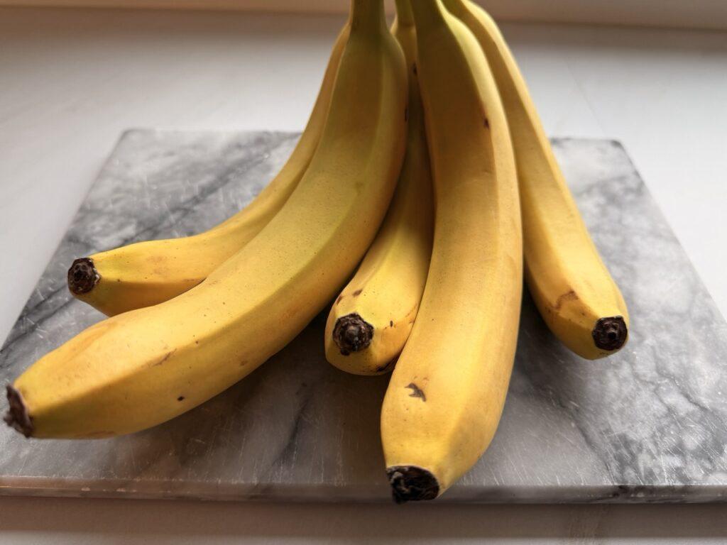 En klase med modne, gule bananer ligger på en marmorfjøl. Bananene har en jevn gul farge med noen små brune prikker og mørke stilker. Lyset fra vinduet gir en naturlig glød til frukten, og den grå marmorfjølen skaper en kontrast til den varme gulfargen. Bananer er fulle av helsefordeler som få kjenner til! Fra bedre søvn til sunn fordøyelse – her er fem gode grunner til å spise en banan hver dag.