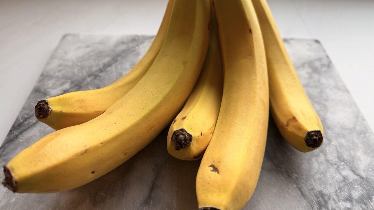 En klase med modne, gule bananer ligger på en marmorfjøl. Bananene har en jevn gul farge med noen små brune prikker og mørke stilker. Lyset fra vinduet gir en naturlig glød til frukten, og den grå marmorfjølen skaper en kontrast til den varme gulfargen. Bananer er fulle av helsefordeler som få kjenner til! Fra bedre søvn til sunn fordøyelse – her er fem gode grunner til å spise en banan hver dag.