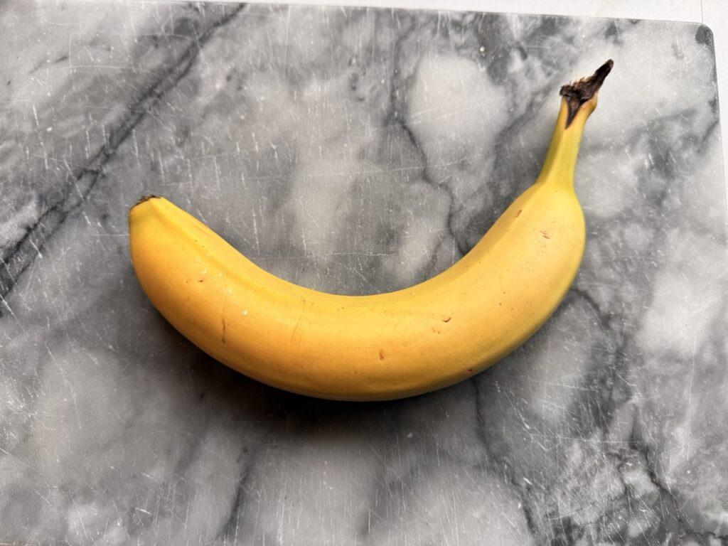 En enkelt, moden banan ligger på en grå marmorfjøl. Bananen har en buet form, jevn gul farge med noen små brune prikker og en mørknet stilk. Marmorfjølen har en naturlig marmorering, som gir et stilrent uttrykk. Bananer er fulle av helsefordeler som få kjenner til! Fra bedre søvn til sunn fordøyelse – her er fem gode grunner til å spise en banan hver dag.