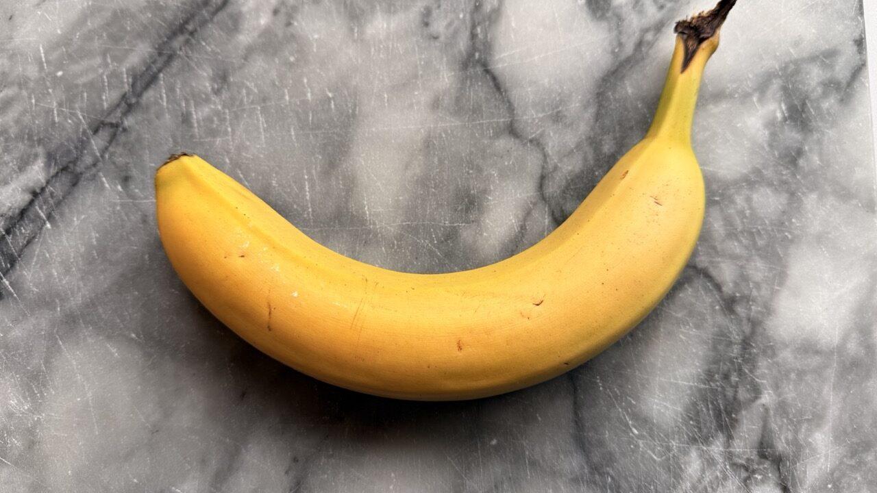 En enkelt, moden banan ligger på en grå marmorfjøl. Bananen har en buet form, jevn gul farge med noen små brune prikker og en mørknet stilk. Marmorfjølen har en naturlig marmorering, som gir et stilrent uttrykk. Bananer er fulle av helsefordeler som få kjenner til! Fra bedre søvn til sunn fordøyelse – her er fem gode grunner til å spise en banan hver dag.