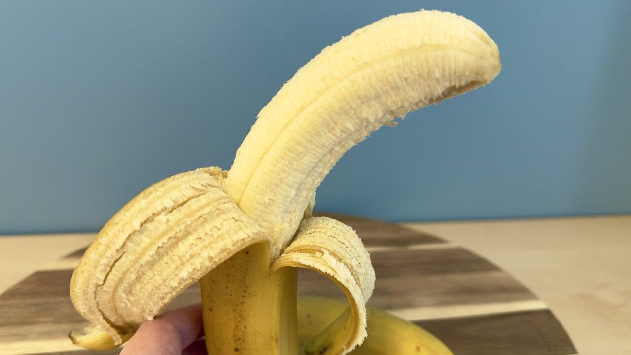 En delvis skrelt banan holdes av en hånd, med den gule skallet brettet tilbake for å avdekke den lyse, modne frukten. I bakgrunnen ligger en hel banan på et rundt skjærebrett av tre, med en blå vegg som bakgrunn.