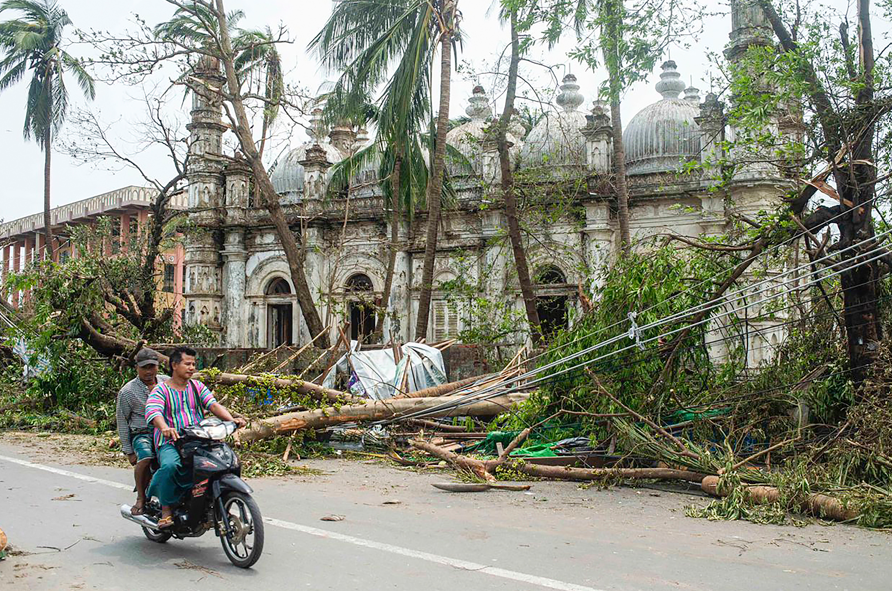 Locals ride motorbike on a road while fallen trees lie near a mosque after Cyclone Mocha in Sittwe township, Rakhine State, Myanmar, Monday, May 15, 2023.