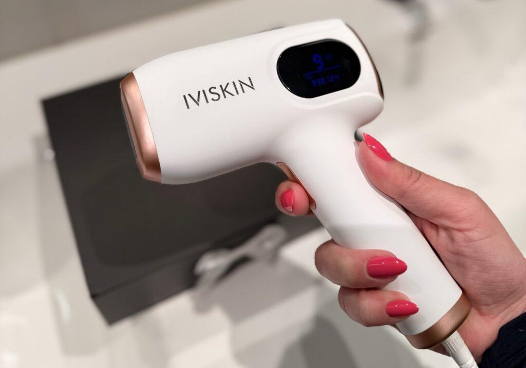 En hånd med lakkerte negler holder Ivyskin G4 IPL-enheten i hvit og roségull, med det digitale displayet synlig på siden. Enheten har pistolgrep og vises foran en uskarp, mørk boks i bakgrunnen.