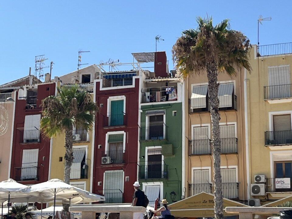 Et sjarmerende gatescene fra en solfylt middelhavsby, Villa Joyosa på Costa Blanca utenfor Alicante i Spania. Fargerike fasader i rødt, grønt, gult og oker står tett i tett under en knallblå himmel. Hver bygning har små balkonger med skodder og tørkesnorer, og flere palmer strekker seg opp foran bygningene. I forgrunnen ser vi folk som rusler, sitter under parasoller og nyter dagen, trolig ved en strandpromenade eller torg. Bildet oser av sørlandsk livsglede, feriestemning og avslappet hverdagsliv i sydlige strøk.
