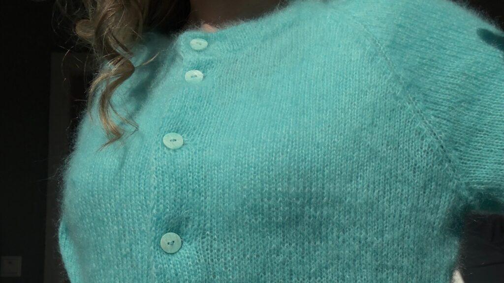 Nærbilde av Marie Cardigan i turkis dobbeltrådet Tynn Silk Mohair. Cardiganen har rund hals, raglanfelling og jevnt knappestolpeparti med tone-i-tone knapper. Garnets glans og fluff gir plagget en lett og eksklusiv struktur.