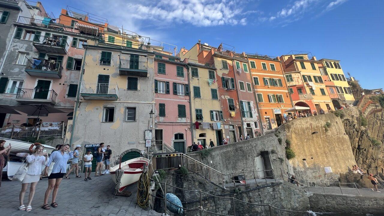 Fargerike hus i Riomaggiore, en av de fem landsbyene i Cinque Terre, Italia. Bildet viser den ikoniske havnen med pastellfargede bygninger, smale smug og turister som nyter utsikten ved sjøen. Riomaggiore er kjent for sin pittoreske beliggenhet, solnedganger og sjarmerende atmosfære langs den italienske rivieraen.