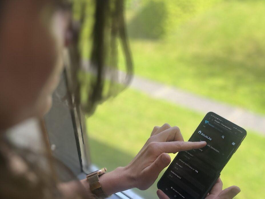 En kvinne står ved et vindu og sjekker boliglånsrenten på mobilen sin. På skjermen vises oversikt over ulike rentebetingelser og boliglånsavtaler. Bildet symboliserer usikkerhet og behov for økonomiske vurderinger i forkant av Norges Banks rentemøte i mai 2025. Innfelt ser vi en portrettbilde av en forbrukerøkonom, som kan være aktuell kilde i saken. Bildet illustrerer temaet: “Rentemøte mai 2025 – dette bør du gjøre nå” og retter seg mot privatpersoner som vurderer grep for å sikre økonomien sin i møte med en uendret eller potensielt endret styringsrente. Avvente, binde renta eller andre tiltak? Dette sier økonomen at du bør gjøre hvis renten holder seg uendret.