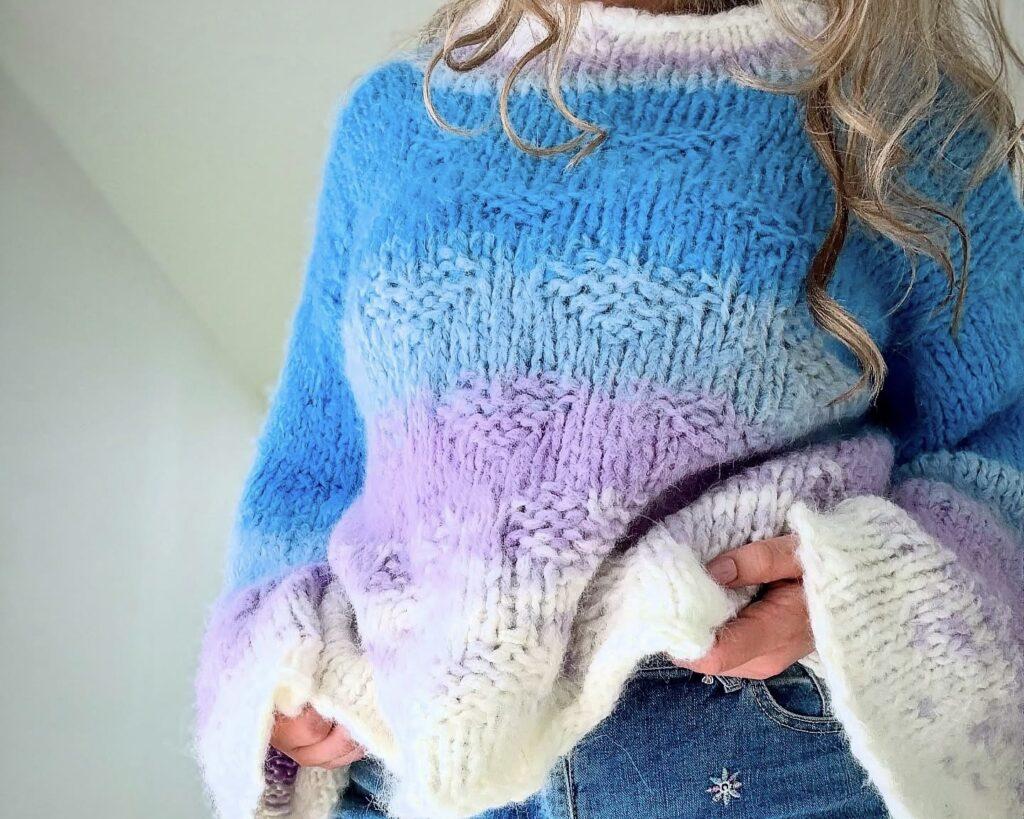 Kvinne iført en håndstrikket, oversized Bryggegenser i pastellfarger – blått, lilla og hvitt – strikket i luftig Camilla Pihl Fnugg garn. Genseren har vide ermer og grov struktur, og styles med jeans med brodert blomst.