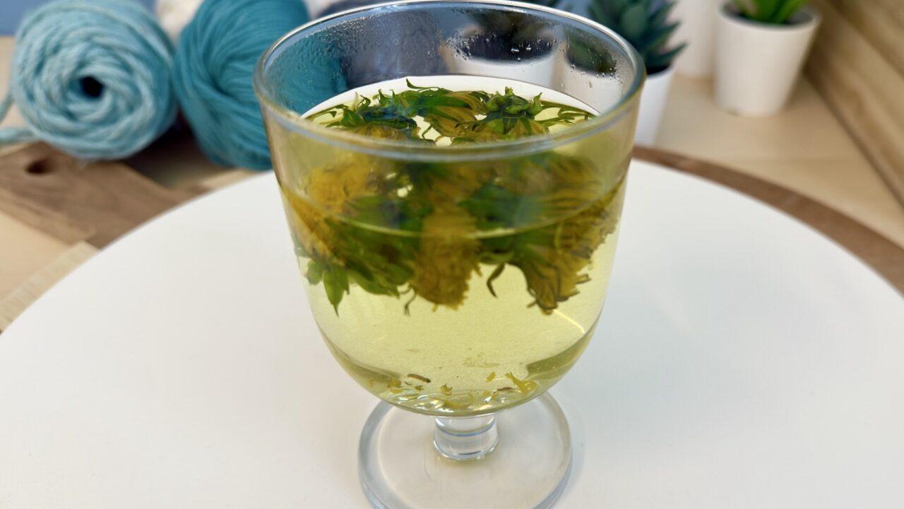 Et glass med gule løvetannblomster trukket i varmt vann for å trekke ut naturlig farge. Væsken har fått en gyllen tone. I bakgrunnen ligger garnnøster og potteplanter, som understreker en kreativ og naturbasert DIY-prosess.
