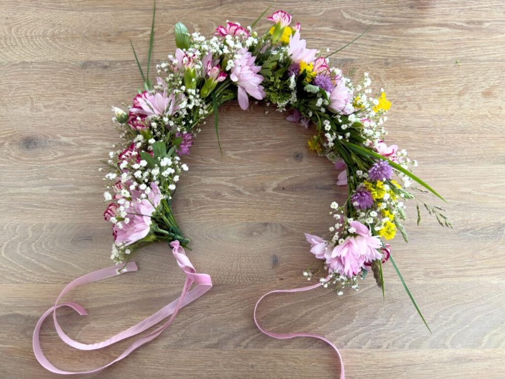 Blomsterkrans laget av rosa, lilla og hvite blomster, med silkebånd i endene. Ligger på trebord. Egnet som hårpynt til midtsommer og sommerfest. Kransen er åpen og knytes i nakken.