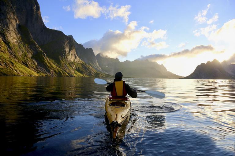 Person i gul kajakk som padler på et rolig blått vann med fjell i bakgrunnen. Her får du de beste tipsene til hvordan du velger riktig kajakk til ditt bruk.