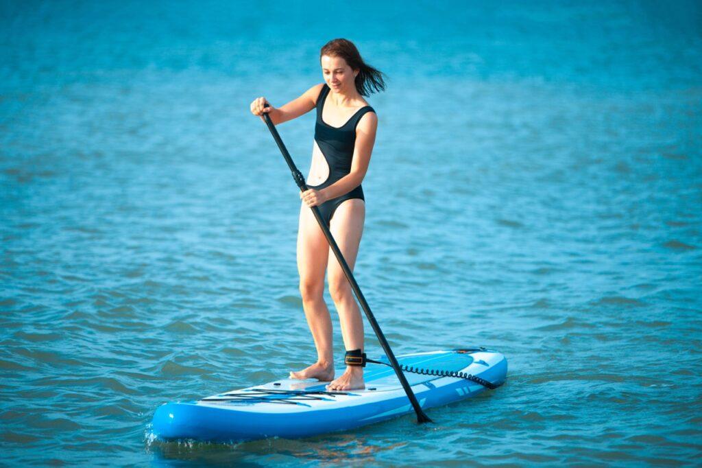 Dame i sort badedrakt som står på et blått SUP-brett, eller paddle board, blått rolig hav. Vi har samlet SUP-brettene som er best i test.